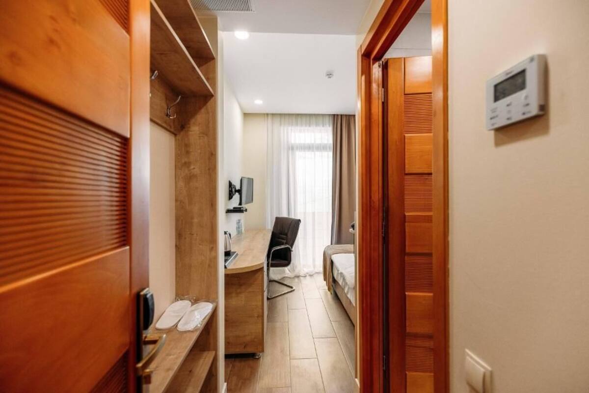 A hotel room with a wooden wardrobe, a desk with a chair, a bed, and a thermostat on the wall.