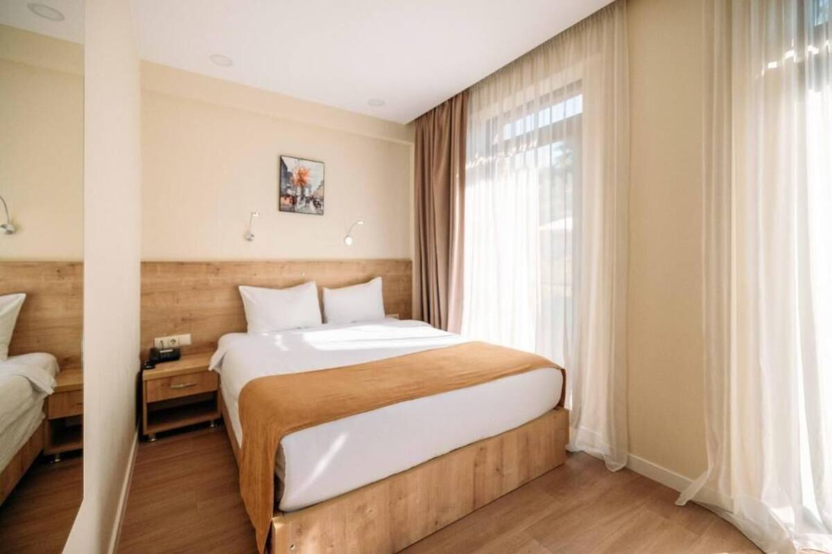 A hotel room with a large bed, a nightstand, a bedside table, a wall-mounted lamp, and a window with sheer curtains.