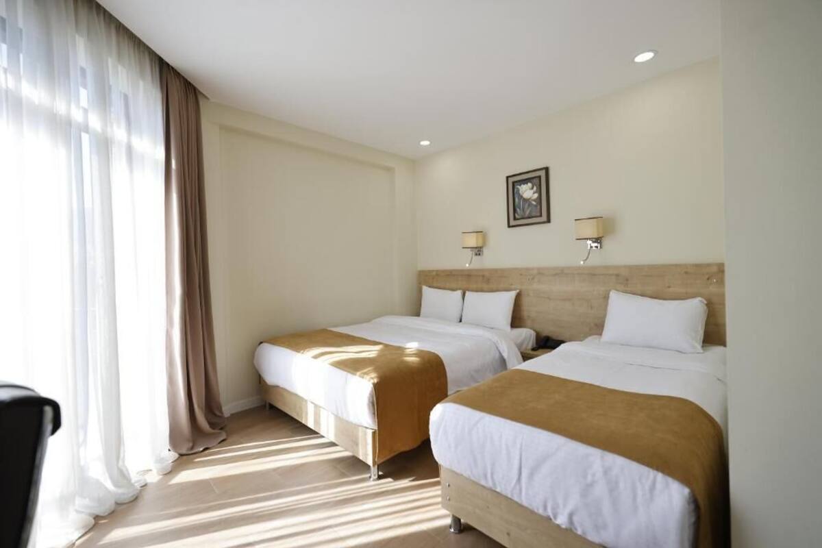 A hotel room with two beds, a wooden headboard, a window with sheer curtains, and a wall-mounted lamp.
