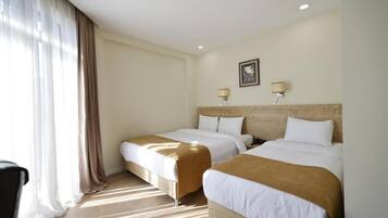 A hotel room with two beds, a wooden headboard, a window with sheer curtains, and a wall-mounted lamp.