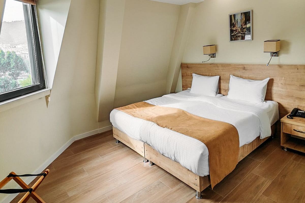 A modern hotel room with a large bed, wooden headboard, and a view of the city through the window.