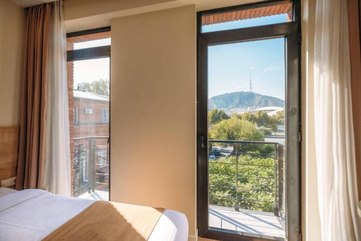 A hotel room with a bed, a window with curtains, and a view of a balcony with greenery and a distant hill.