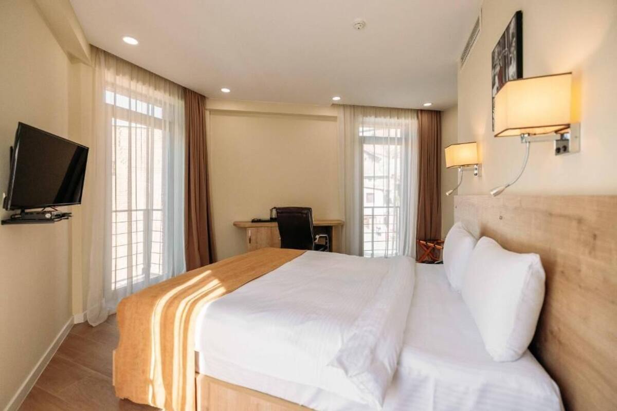 A hotel room with a large bed, a desk, a chair, a television, and two wall-mounted lamps.