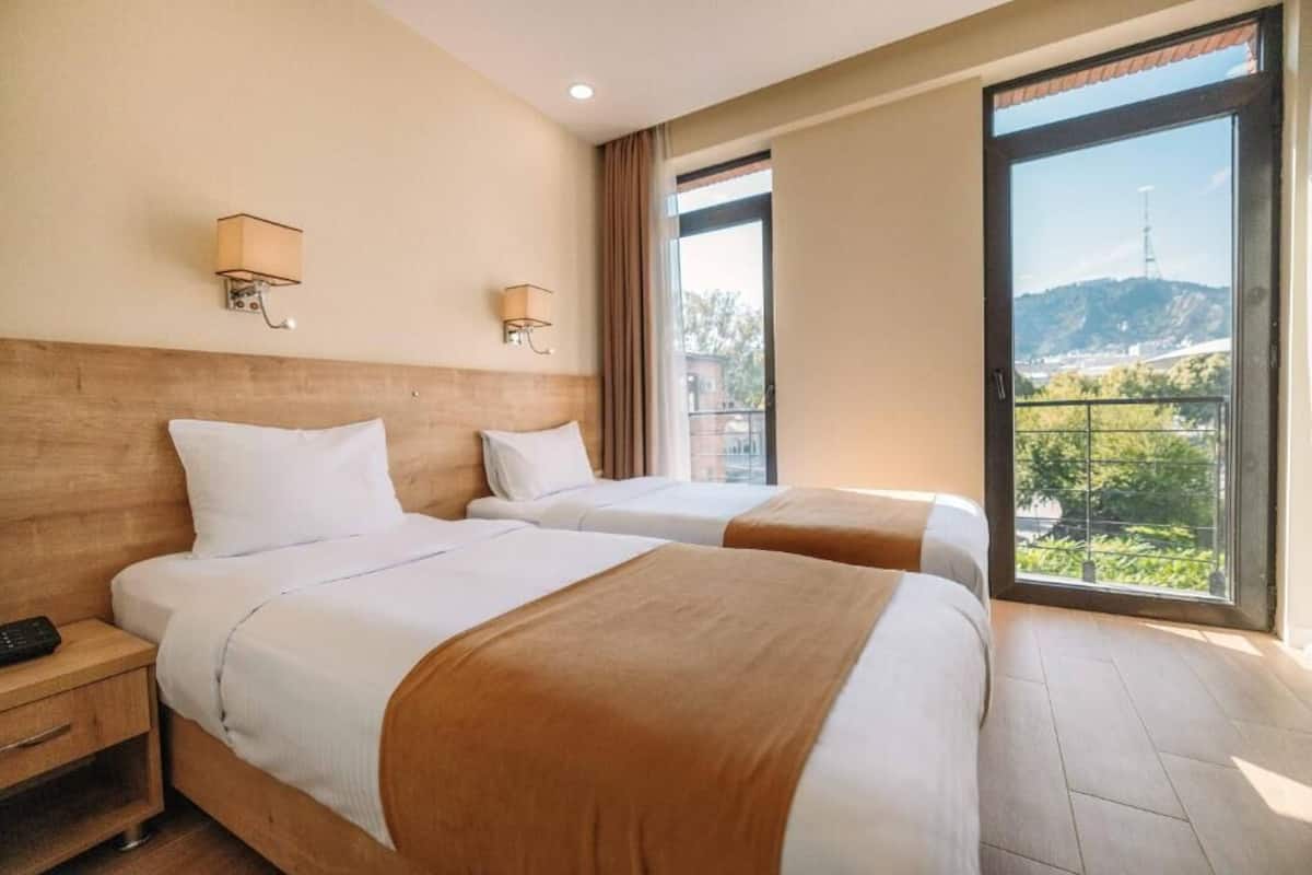 A hotel room with two beds, a nightstand, and a large window offering a view of a hill and a tower.