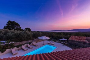 A pool area with lounge chairs and umbrellas, surrounded by stone walls and vegetation, with a view of distant hills and a sunset sky. - Holiday house LUNA with pool (Bajčići)