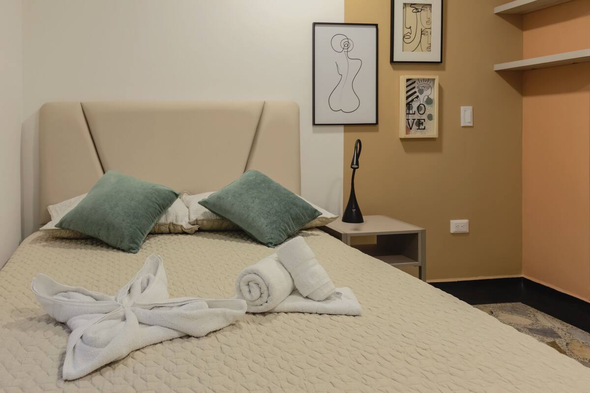 Habitació d'hotel amb un llit, dos coixins, una taula de nit i una paret amb quadres.