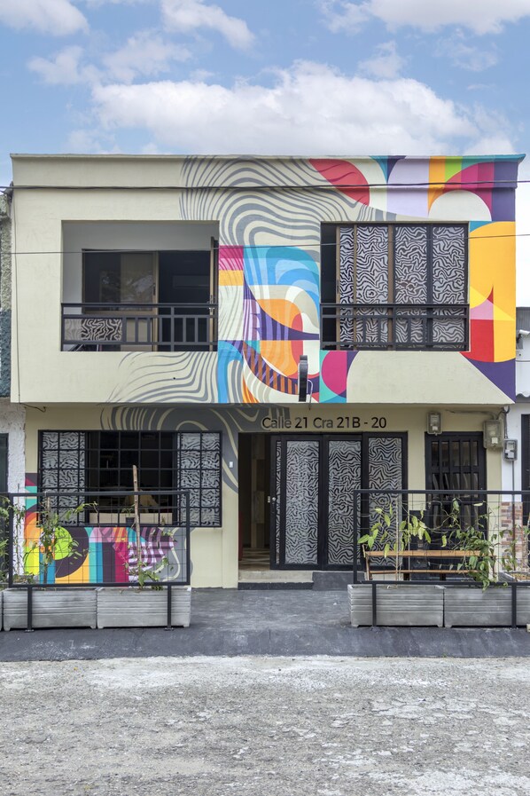 Un edificio de dos pisos con un mural colorido en la fachada, un balcón y un cartel que indica 