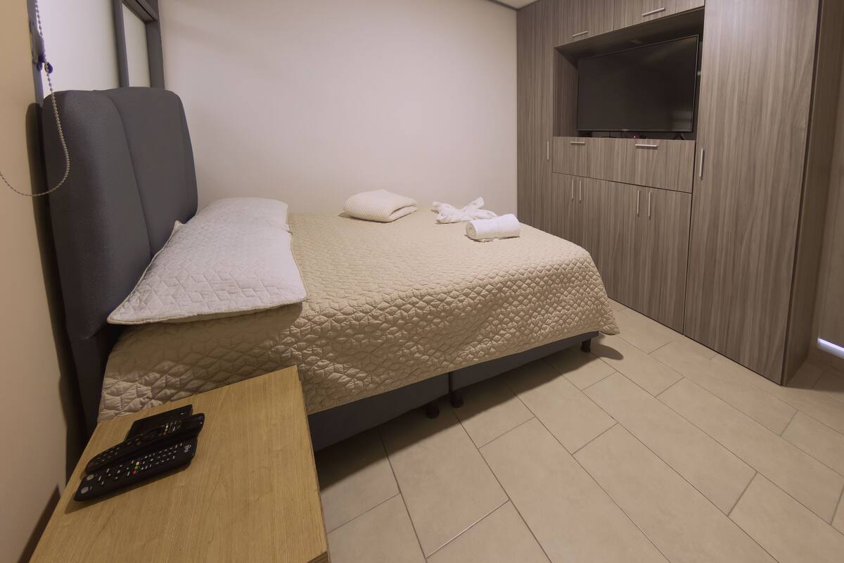 Un dormitori amb un llit, un televisor fixat a la paret i una petita taula de nit de fusta.