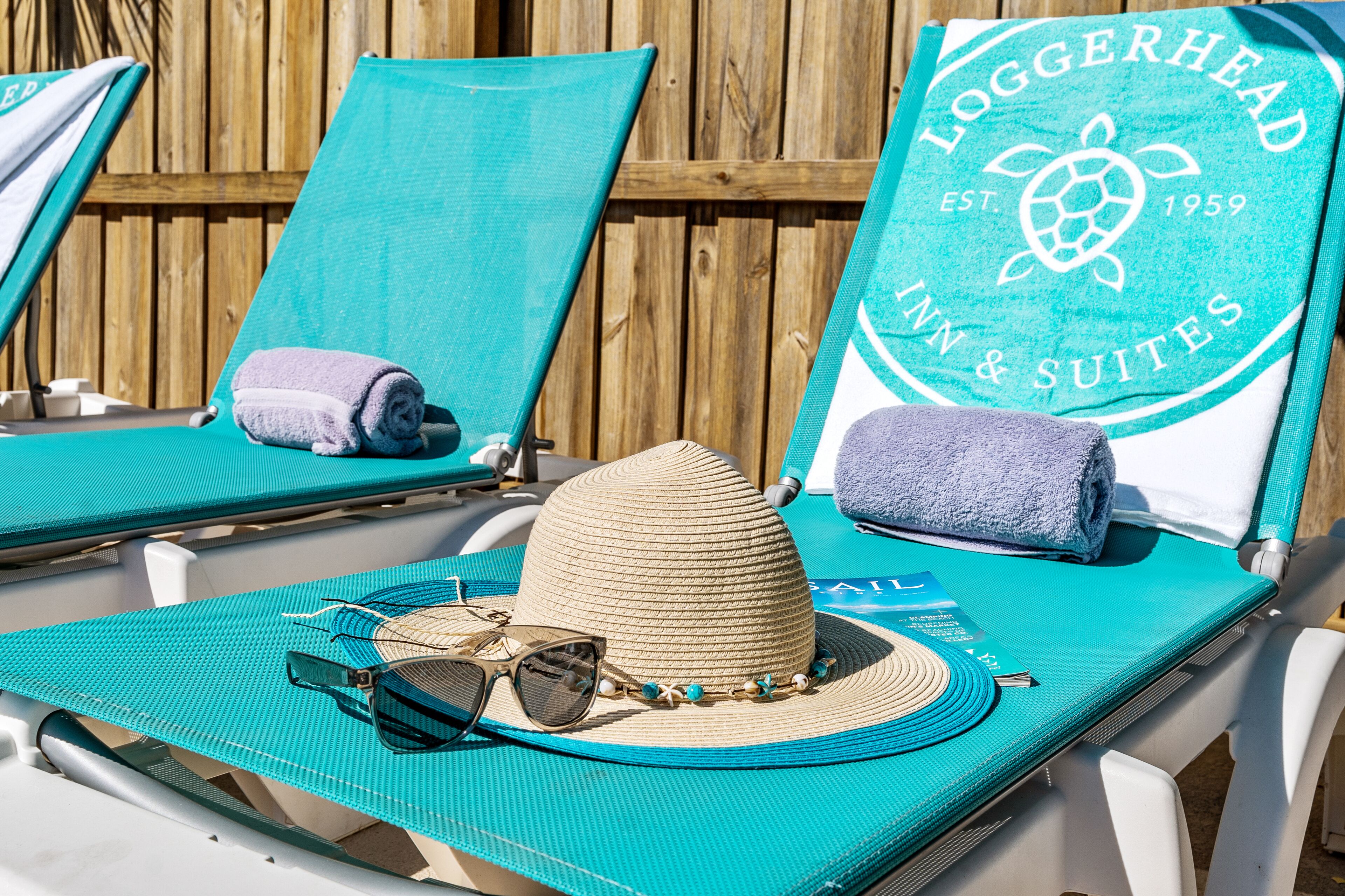Un'area relax in spiaggia con poltrone color turchese, un cappello di paglia, occhiali da sole e un asciugamano. Un cartello recita: "Loggerhead Inn & Suites".