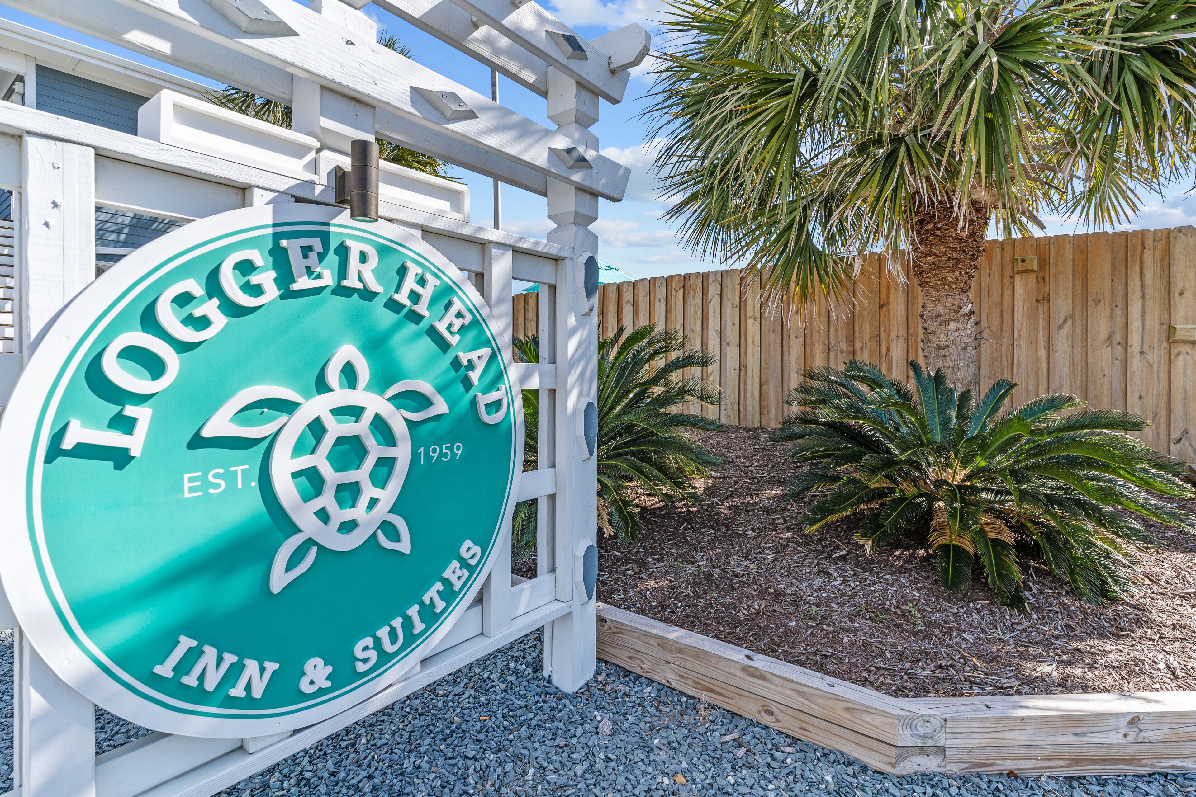A sign for Loggerhead Inn & Suites with a turtle logo, established in 1959, surrounded by a wooden fence and palm trees.