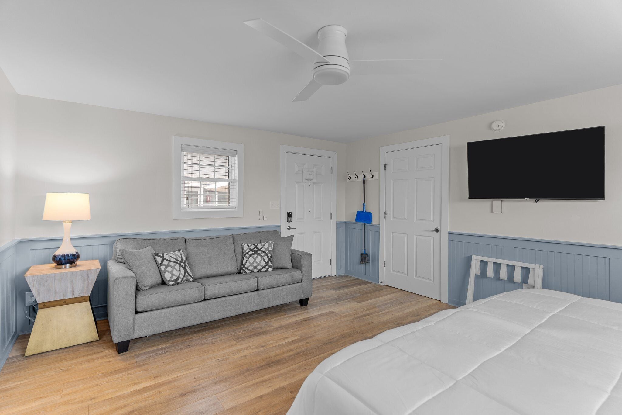 A bedroom with a bed, a grey sofa, a wooden nightstand, a wall-mounted TV, and a ceiling fan.