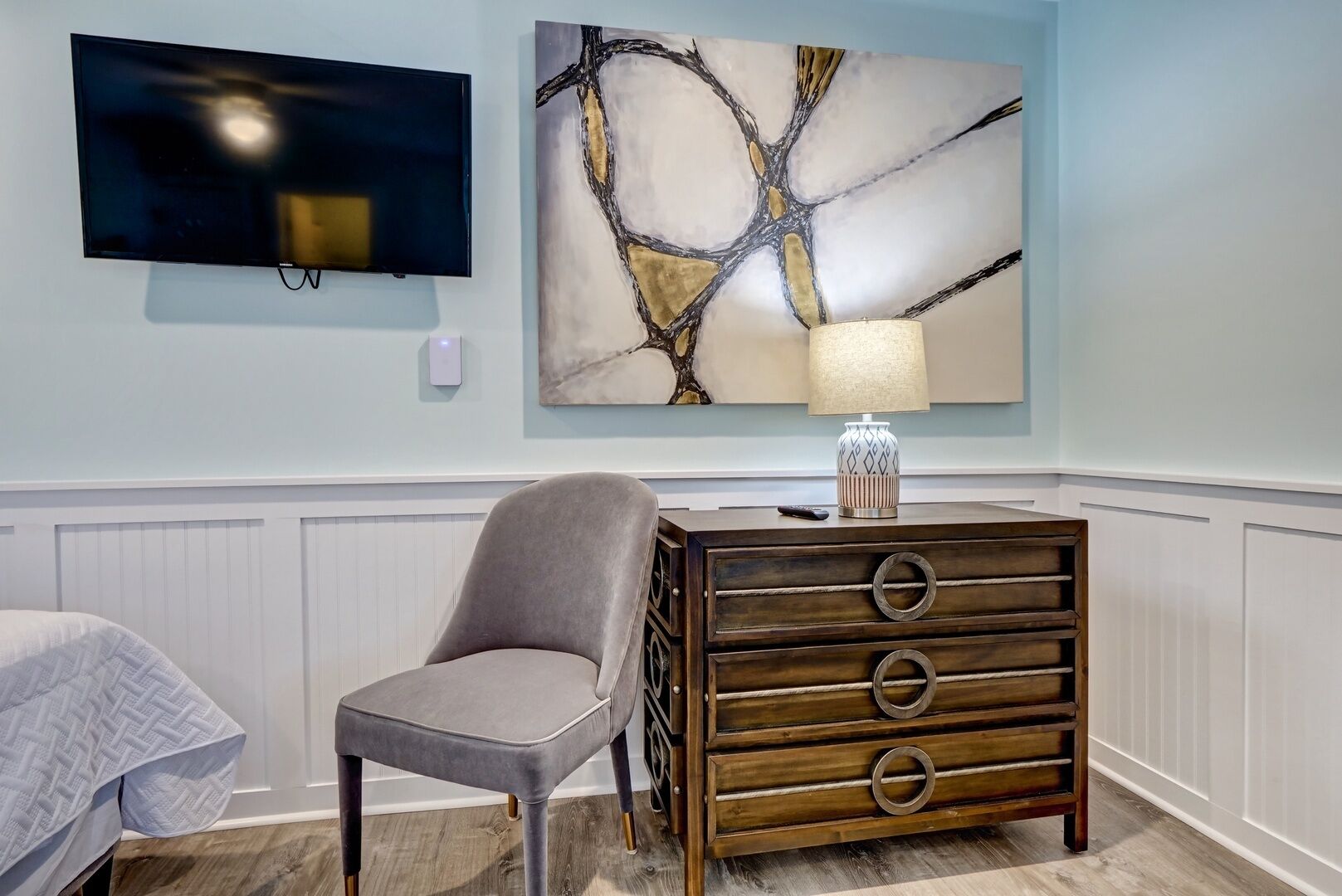 A room with a bed, a gray armchair, a wooden dresser with drawers, a wall-mounted TV, and a large abstract painting.