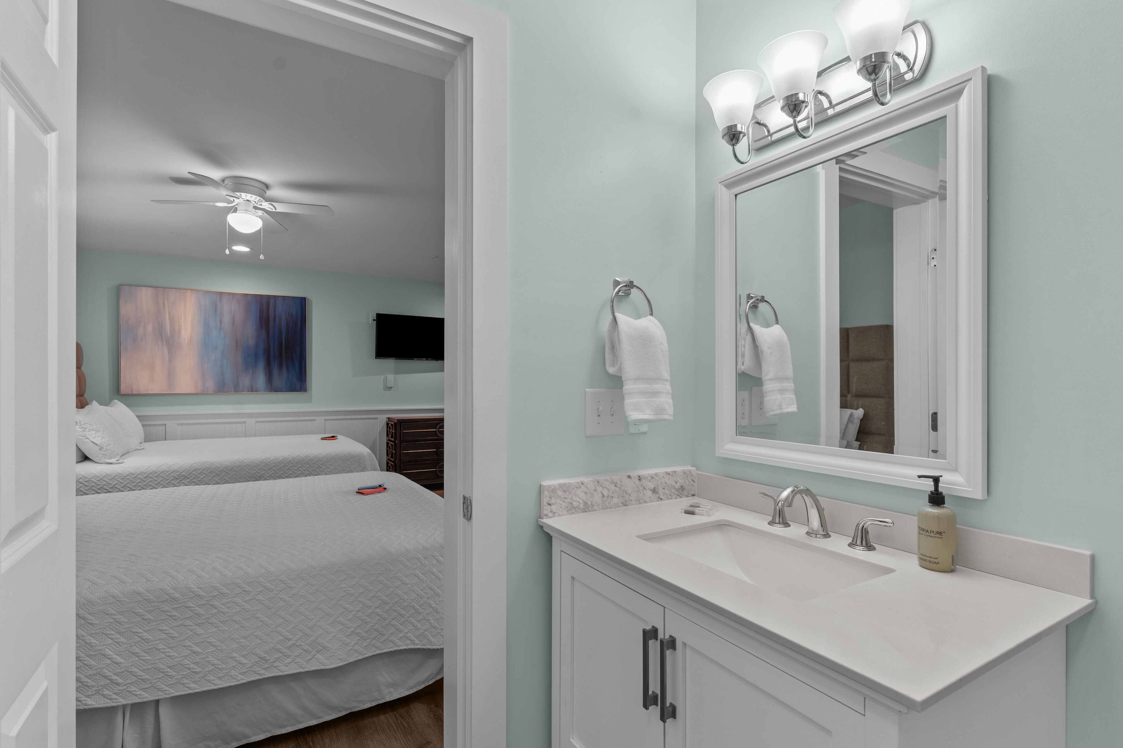 Une chambre d’hôtel avec deux lits, une salle de bain équipée d’un lavabo et d’un miroir, et un ventilateur de plafond.