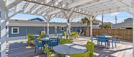 An outdoor patio with wooden deck, white pergola, and outdoor seating with chairs and tables.