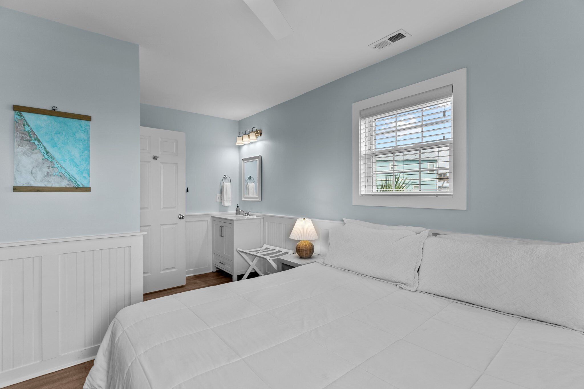 A bedroom with a bed, a nightstand, a lamp, a window with blinds, a door, and a wall-mounted picture.