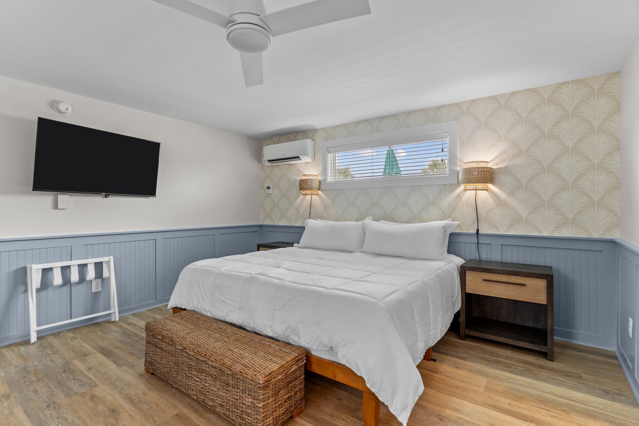 Une chambre à coucher comprenant un lit, une table de chevet, un téléviseur, un appareil de climatisation et une fenêtre avec des stores.