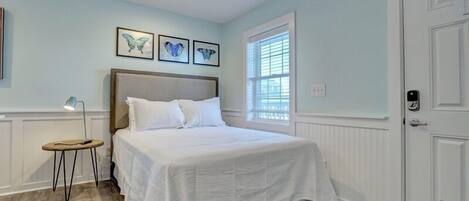 A bedroom with a large bed, a small stool, a window with blinds, and a door.