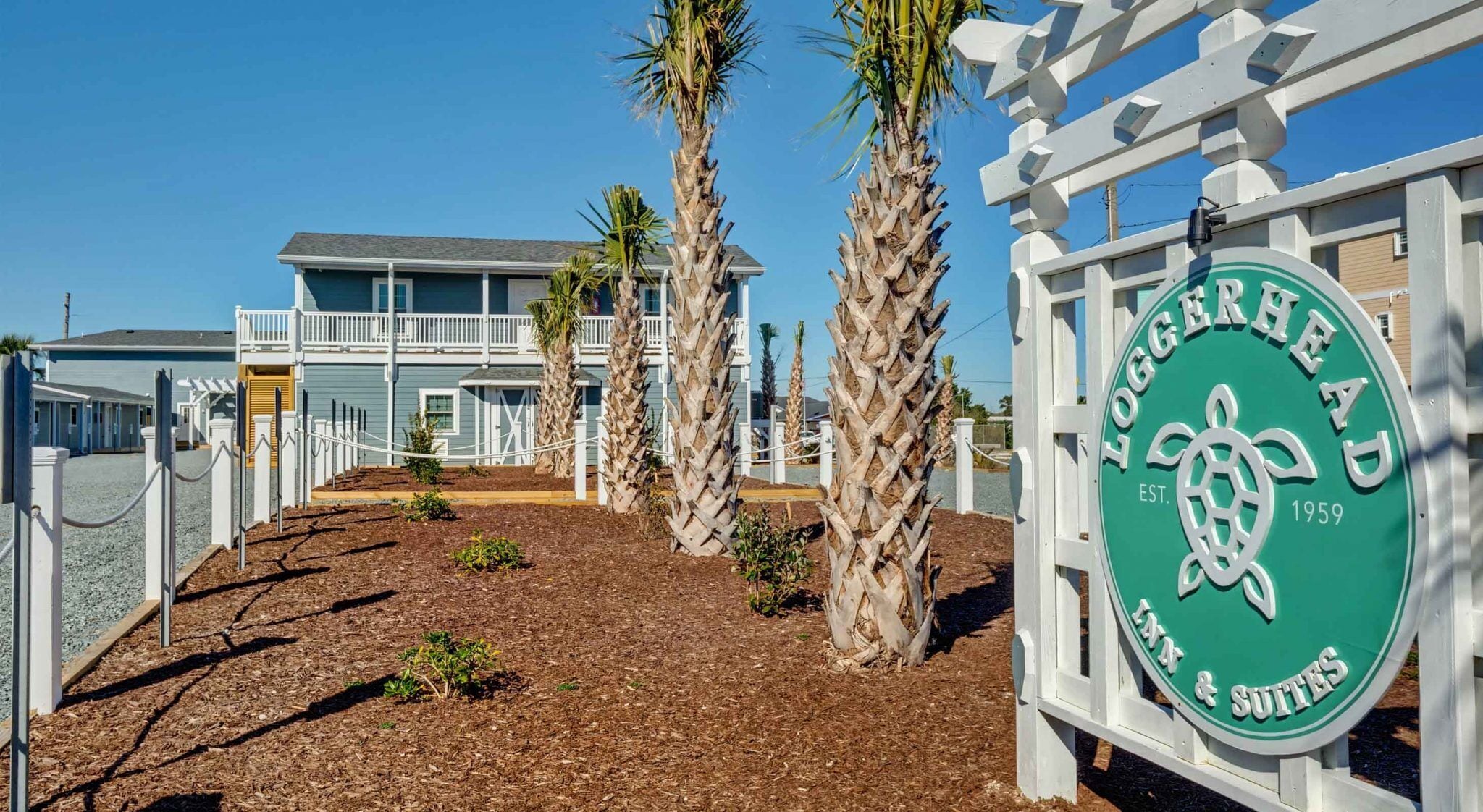 A beachfront property with a sign reading "Loggervhead & Suites" established in 1959, featuring a two-story building with a balcony and palm trees in the foreground.