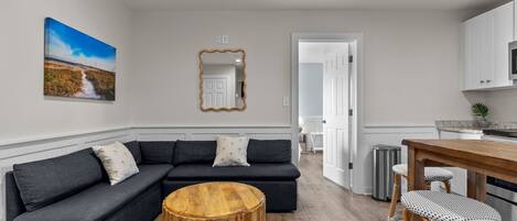 Una sala de estar moderna con un sofá seccional gris oscuro, una mesa de centro redonda de madera y un comedor con mesa de madera y taburetes de mimbre.