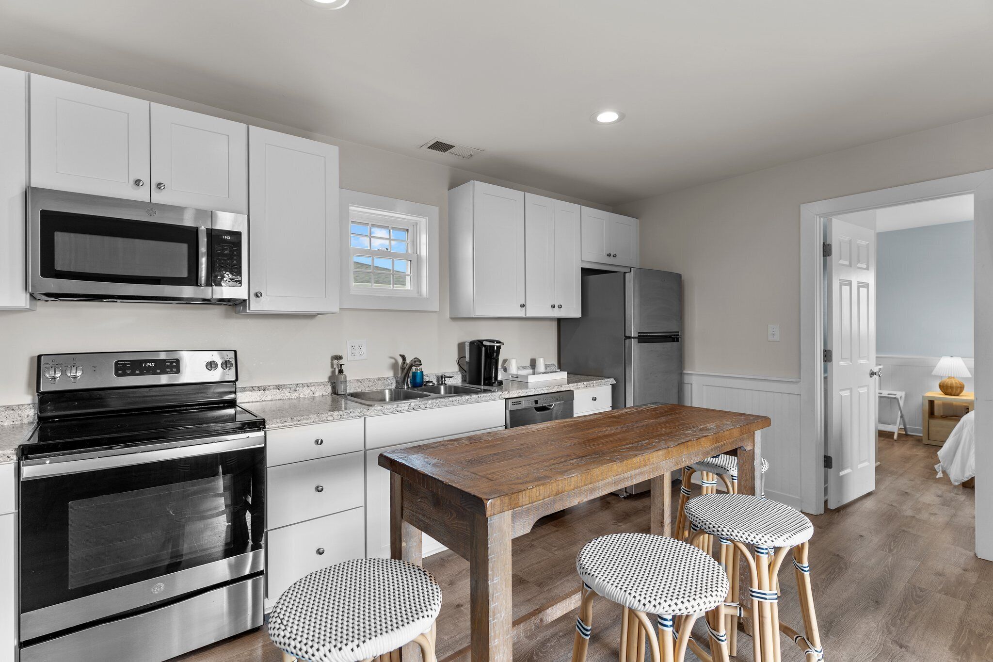 A modern kitchen with white cabinetry, stainless steel appliances, a wooden table, and two wicker stools.