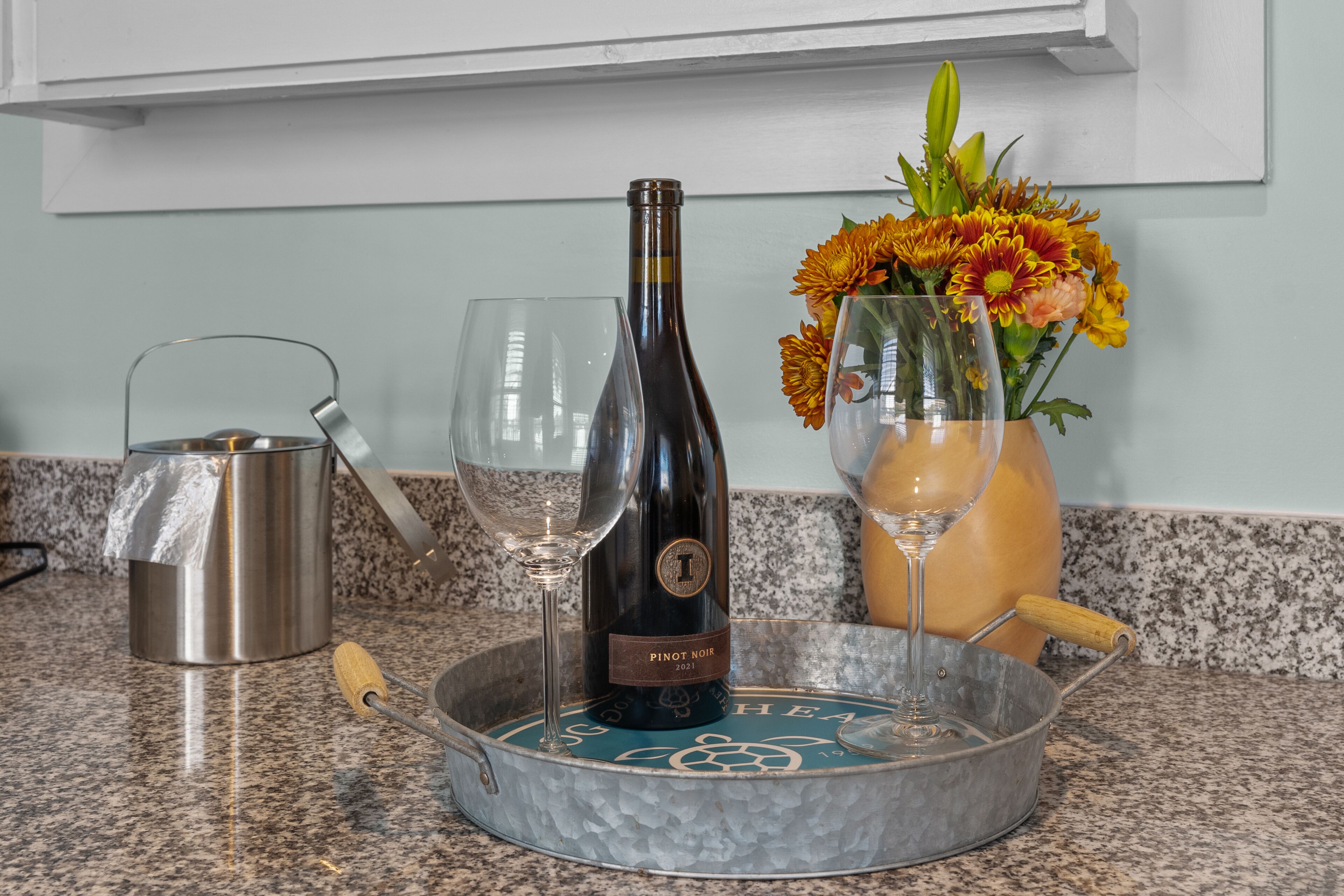 A kitchen counter with a wine bottle, a glass, a metal pitcher, and a vase with flowers.