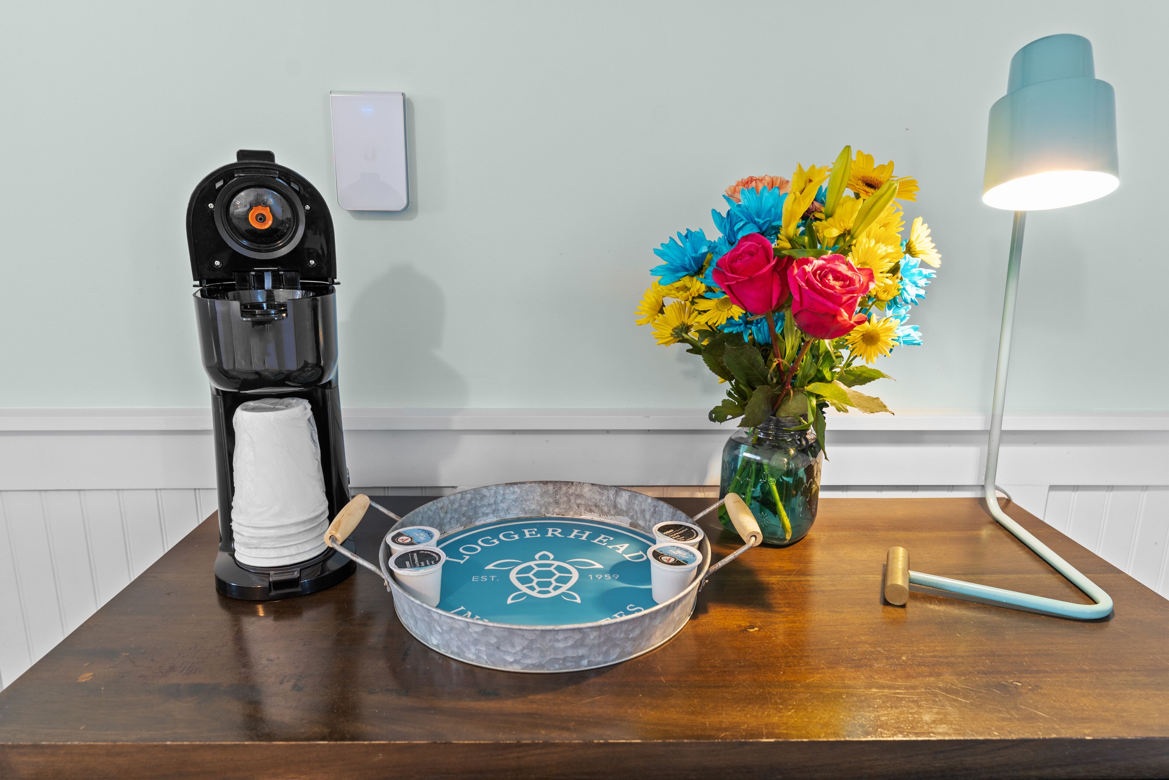 A wooden table with a coffee maker, a tray with a logo, a vase with flowers, and a lamp.