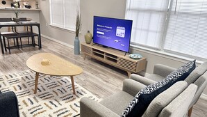 Moderni olohuone, jossa on sohva, sohvapöytä ja televisio. Huoneessa on myös ruokailutila, jossa on tuoleja ja pöytä. Lattiassa on puulattia ja siinä on kuvioitu matto.