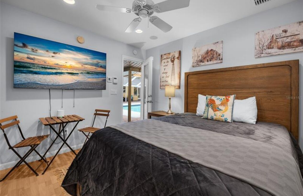A bedroom with a large bed, a ceiling fan, a painting of a sunset, and a small table with chairs.