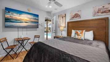 A bedroom with a large bed, a ceiling fan, a painting of a sunset, and a small table with chairs.