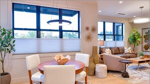 A modern living room with a round wooden table, white chairs, a sofa, and a coffee table. There are large windows with blinds, a hanging light fixture, and decorative wall art.