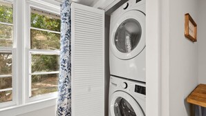 A laundry room with two stacked washing machines, a window with a view of greenery, and a wooden shelf with a framed item. - Downtown Dahlonega Destination - Dog Friendly (Dahlonega)