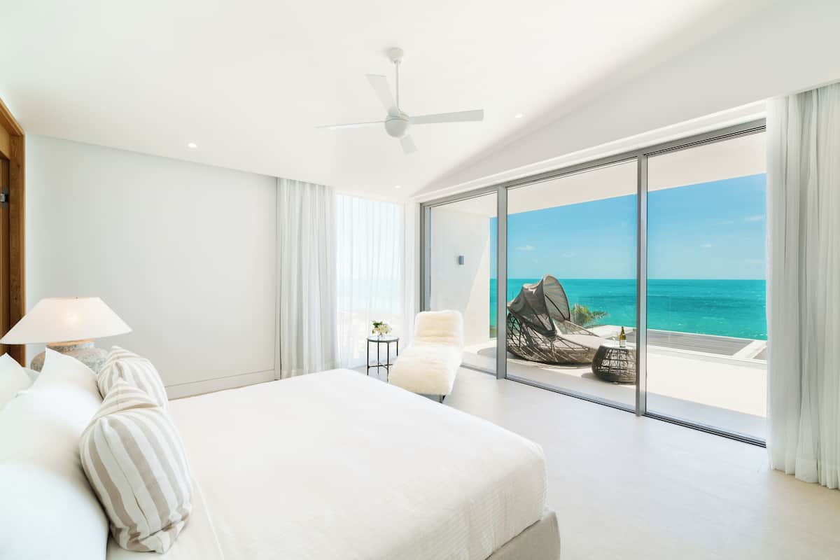 A bedroom with a large bed, a ceiling fan, and a view of the ocean through floor-to-ceiling glass doors.
