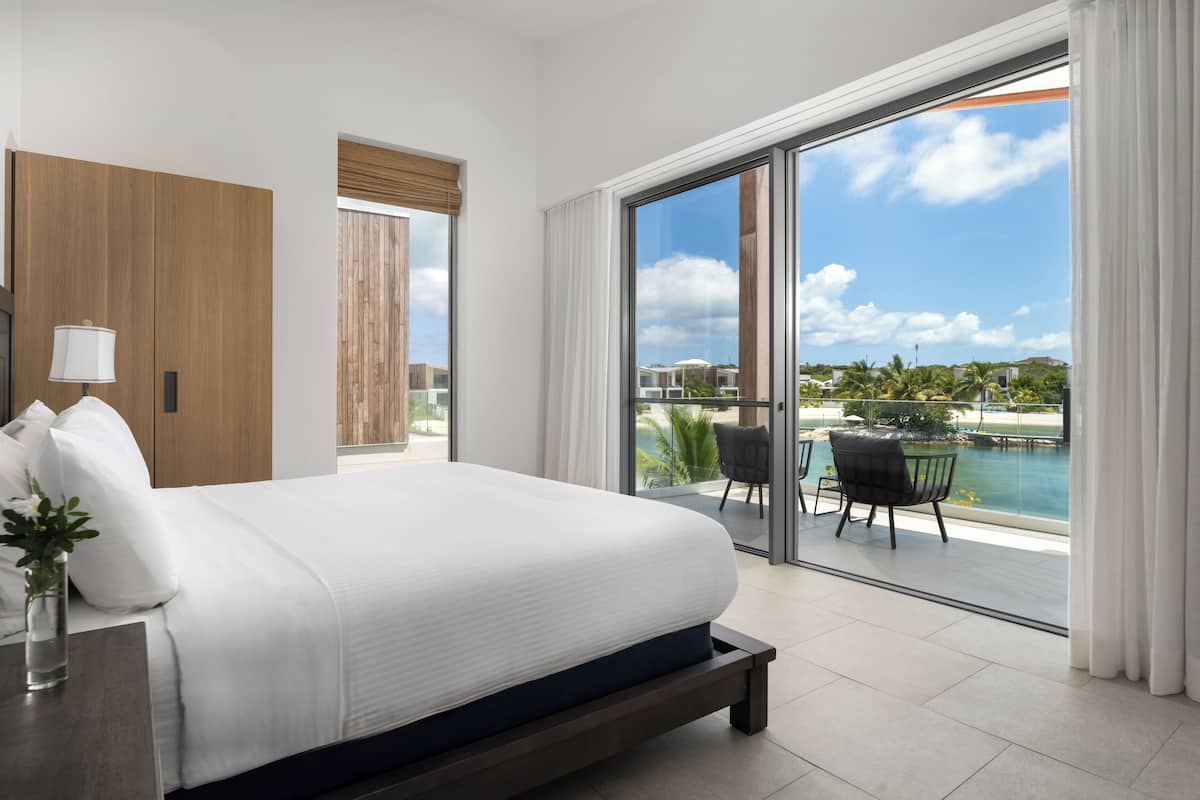A modern hotel room with a large bed, a nightstand, a lamp, a view of the pool and palm trees through a sliding glass door.