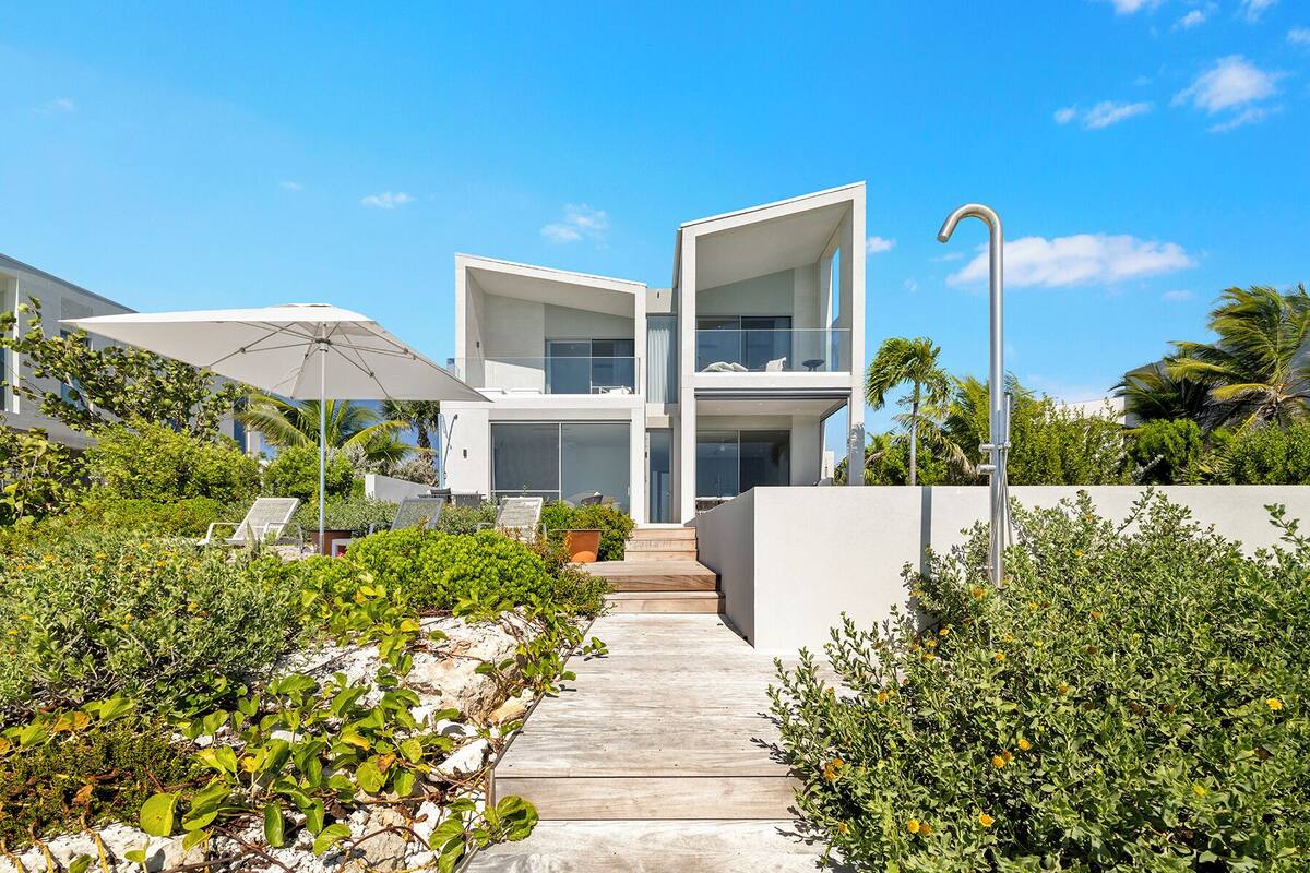 A modern two-story house with a concrete pathway leading to the entrance, surrounded by lush greenery and palm trees.