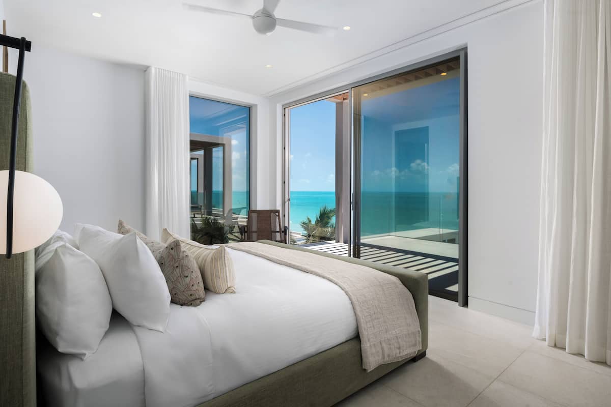 A modern bedroom with a large bed, a ceiling fan, and a view of the ocean through a sliding glass door.