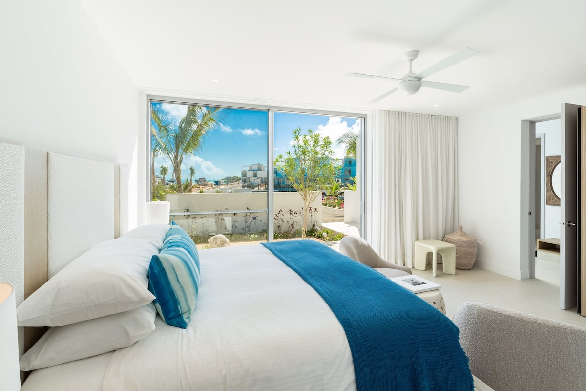 A modern bedroom with a large bed, a ceiling fan, and a view of palm trees and buildings outside.