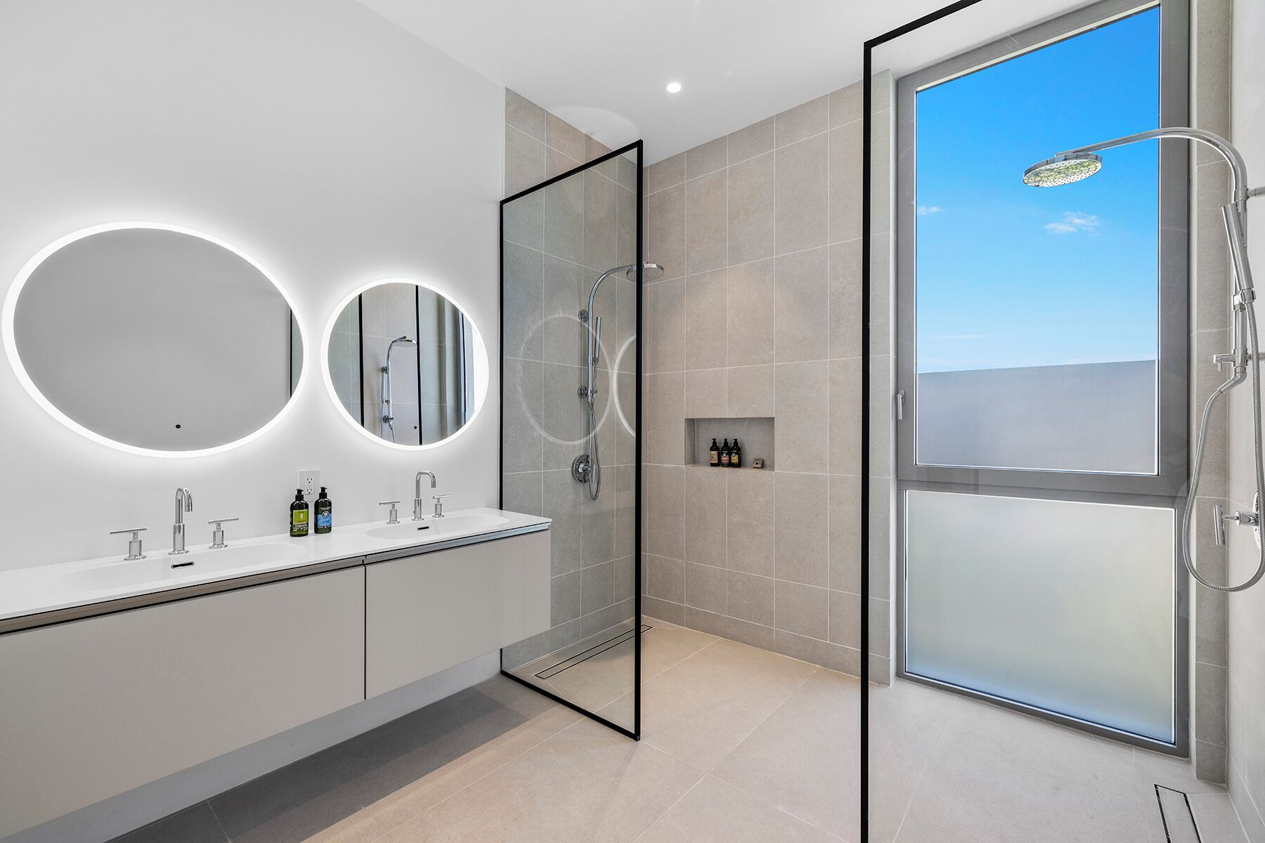 A modern bathroom with a glass shower enclosure, two round mirrors, and a built-in shelf with toiletries.