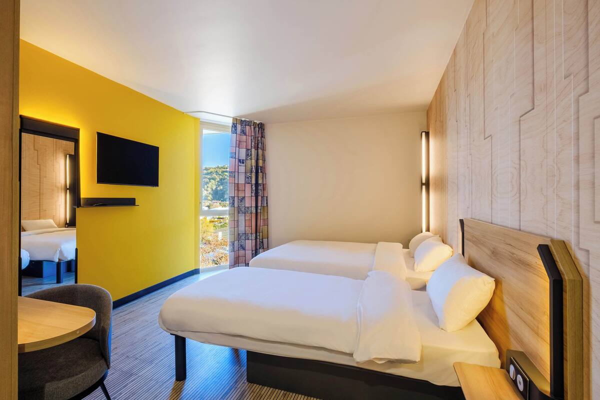 A hotel room with two beds, a TV mounted on the wall, a small table, and a view of the outdoors.