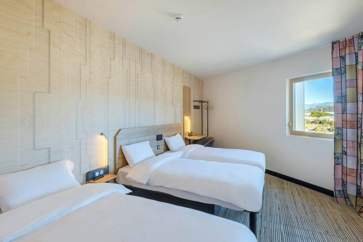 A hotel room with three beds, a wooden headboard, a nightstand with a lamp, and a window with a view of the landscape.