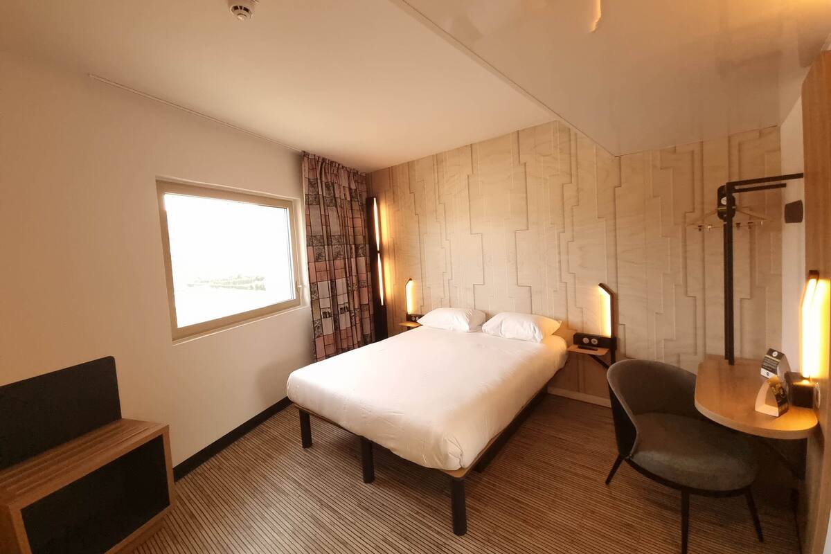 A hotel room with a large bed, a desk with a chair, a window with curtains, and a wall-mounted lamp.