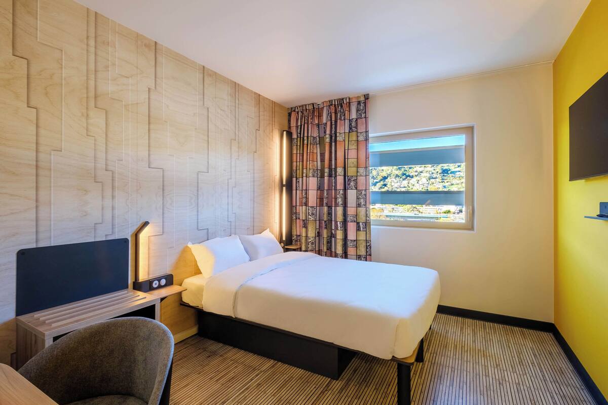 A hotel room with a bed, a desk with a flat-screen TV, a chair, a nightstand with a lamp, and a window with a city view.