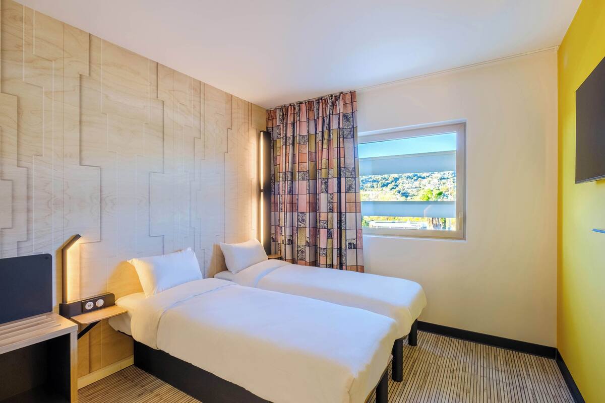 A hotel room with two beds, a TV, a desk, and a window with a view.