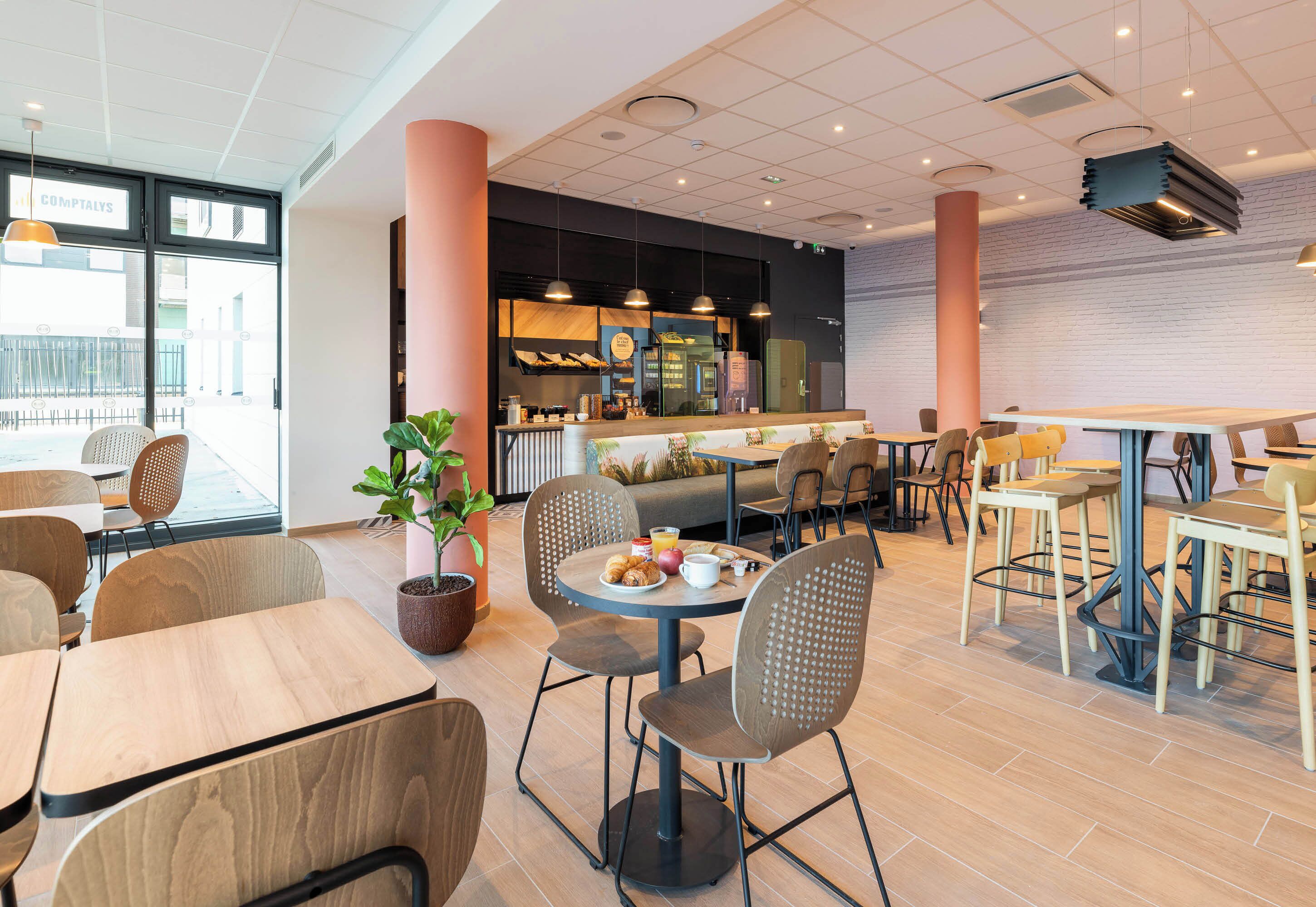 Ein modernes Café mit Holztischen und -stühlen, einem zentralen Sitzbereich und einer Küche im Hintergrund.