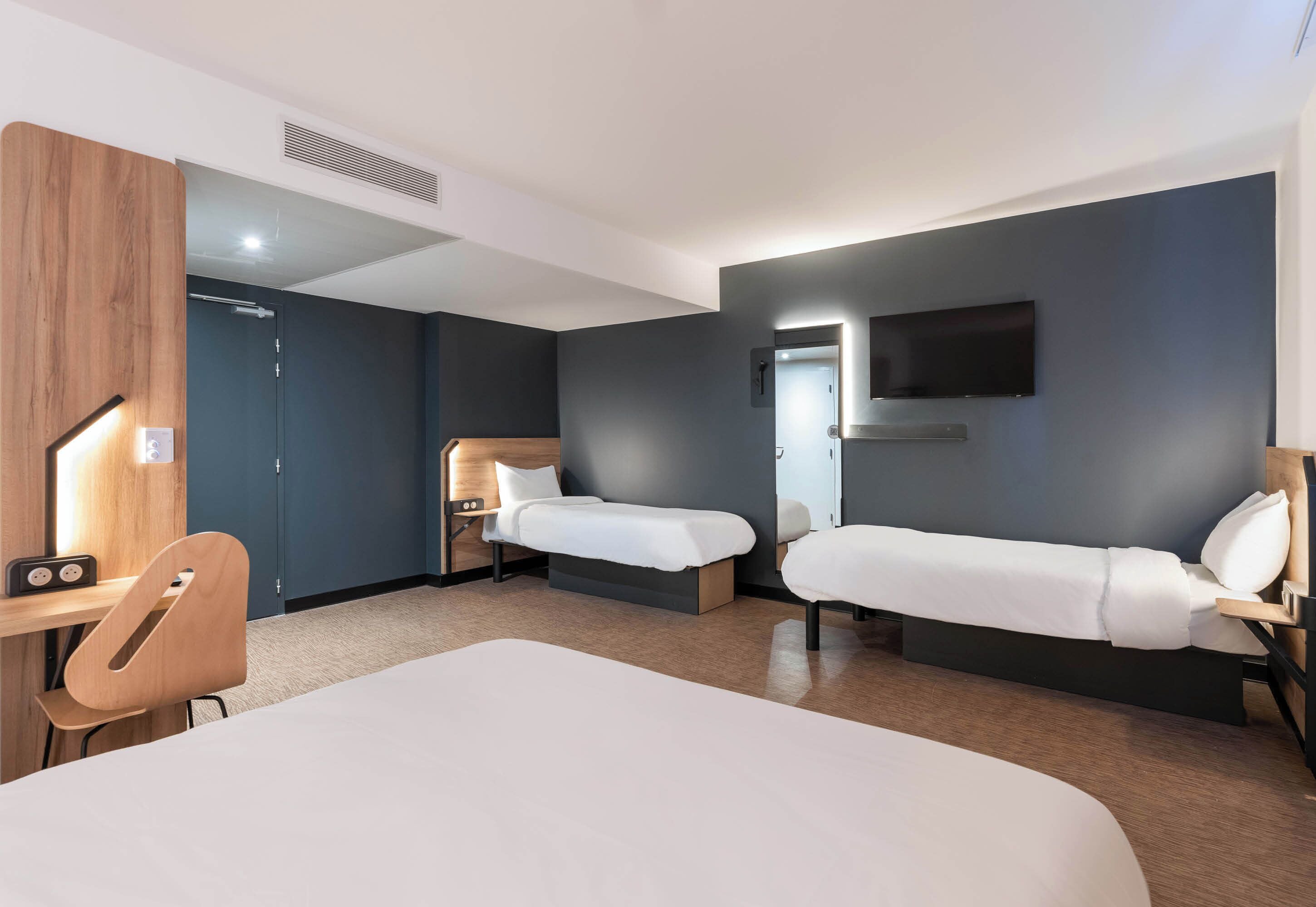 Ein Hotelzimmer mit zwei Einzelbetten, einem Schreibtisch mit Stuhl, einem an der Wand befestigten Fernseher und einer Holzvertäfelung an der linken Wand.