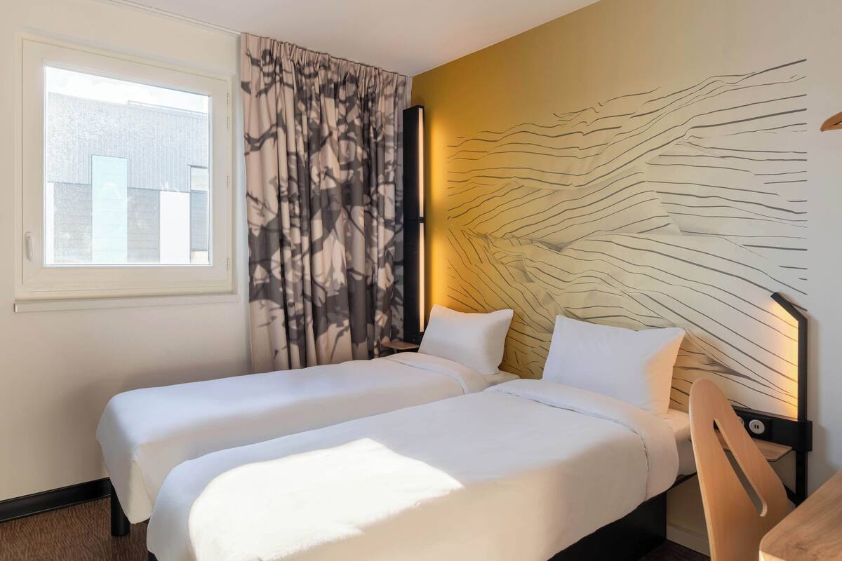 Une chambre d’hôtel avec deux lits, une grande fenêtre, un luminaire fixé au mur et une chaise.