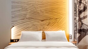 A bed with white bedding, a headboard with a wavy design, and a bedside table with a lamp. - B&B HOTEL Massieux Genay (Genay)