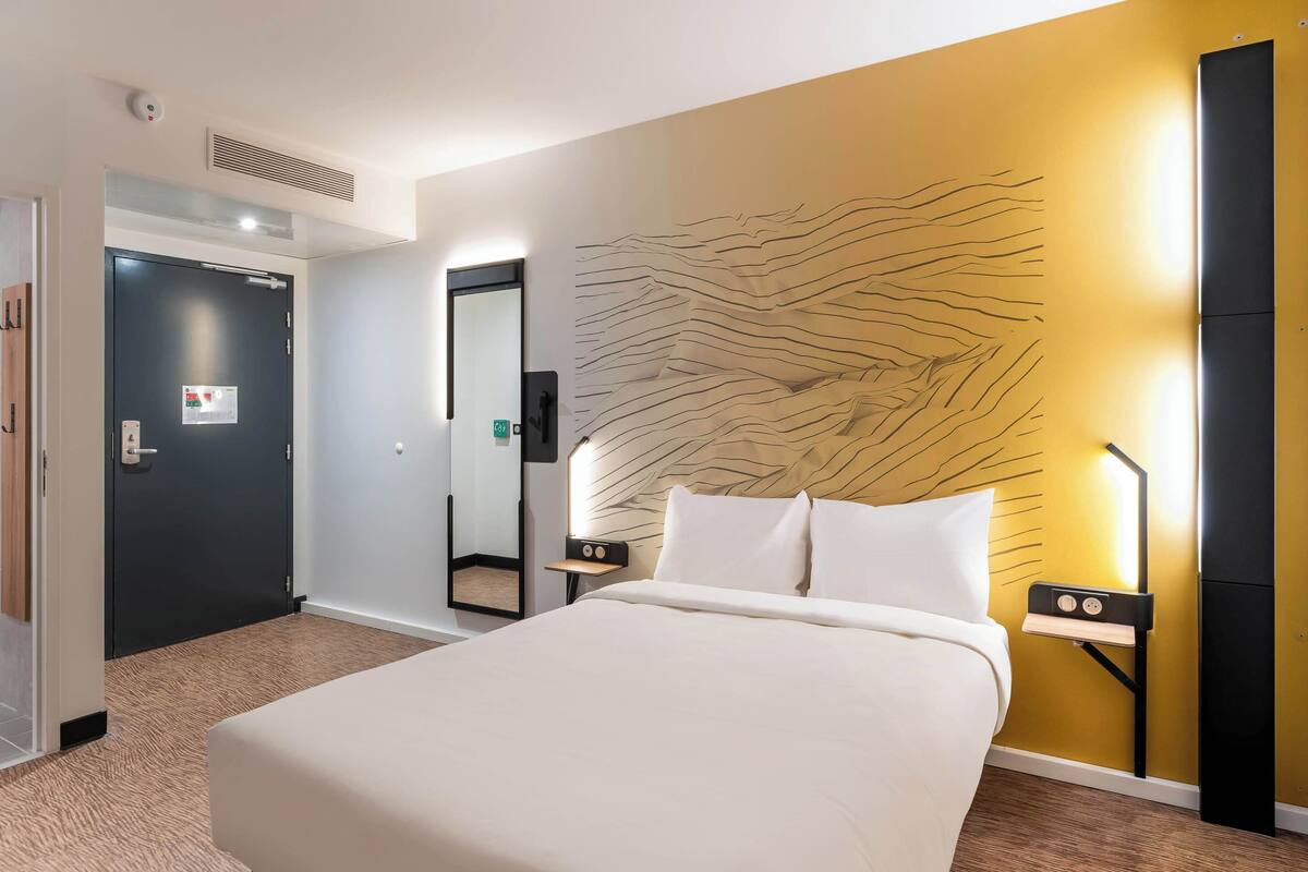Une chambre d’hôtel avec un lit, une table de chevet avec une lampe, une œuvre d’art accrochée au mur et une porte avec une pancarte.