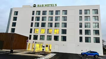 A B&B hotel with a modern facade, multiple windows, and a parking area with a blue car.