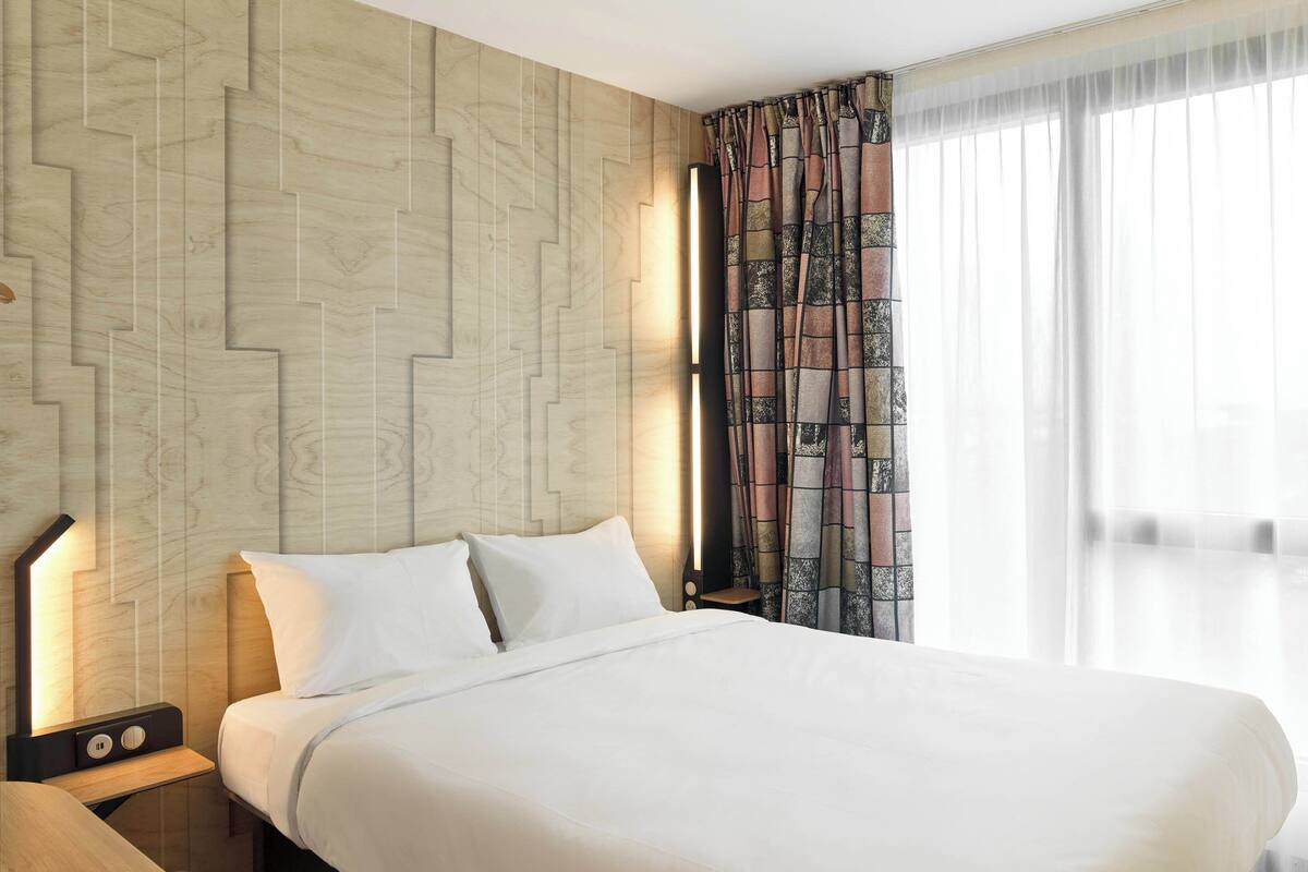 Une chambre d’hôtel moderne avec un grand lit, une table de chevet avec une lampe et une vue sur la ville à travers des rideaux transparents.