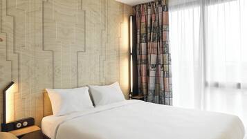 Habitación de hotel moderna con una cama grande, una mesita de noche con lámpara y vistas a la ciudad a través de cortinas translúcidas.